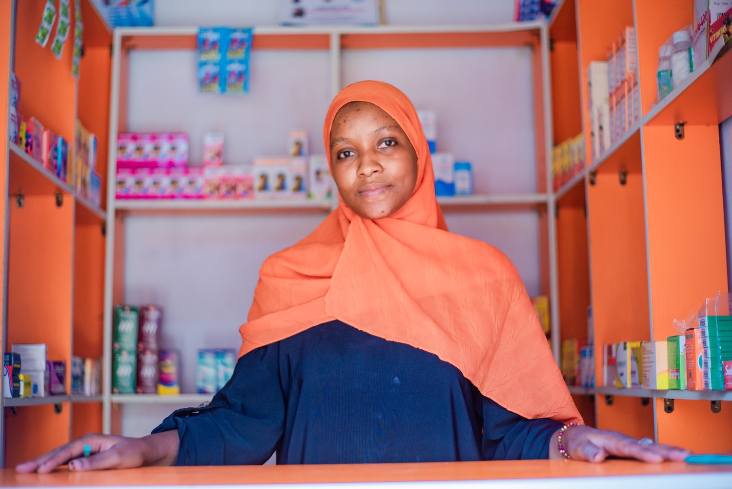 A community pharmacy worker stands behind a counter stocked with branded health commodities