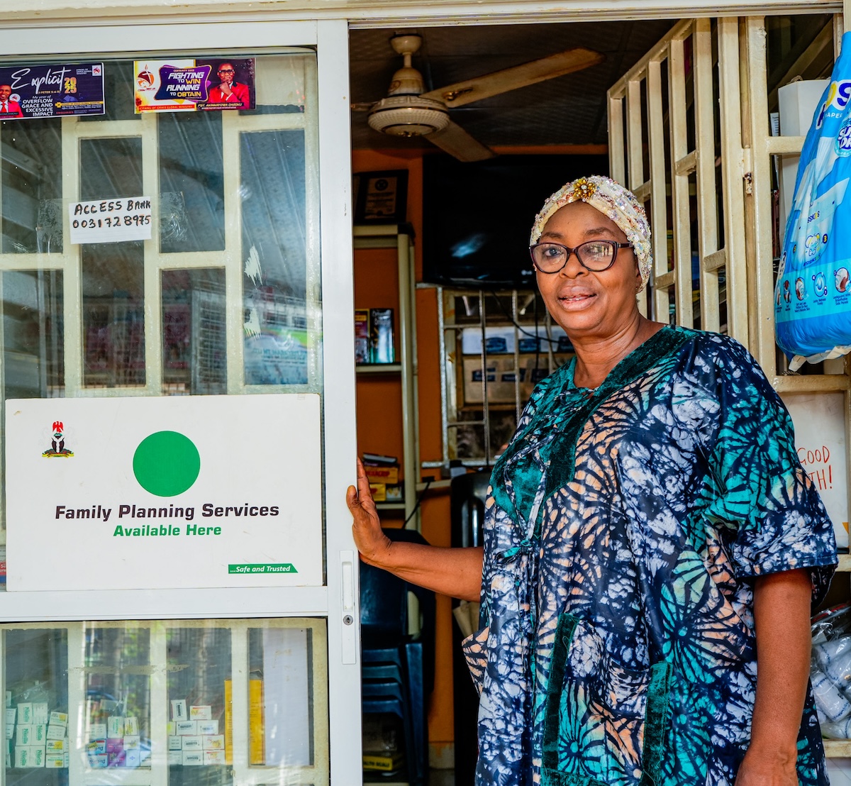 A community pharmacy proprietor stands beside a branded 'Family Planning Services Available Here' sign — one of the outlets through which SFH delivers its family planning programme.