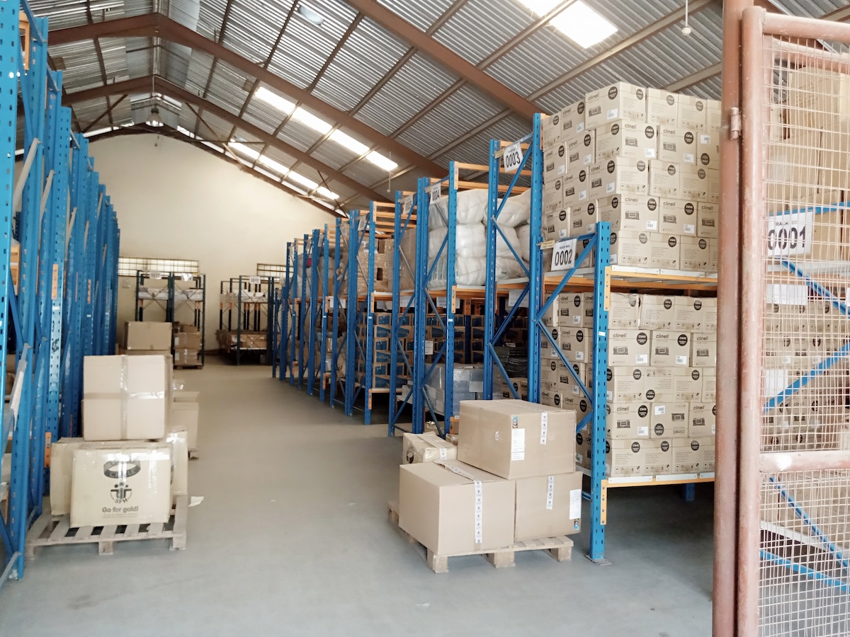 SFH Access warehouse — pallets of boxed health commodities stacked on blue racking under a corrugated roof, ready for last-mile distribution.